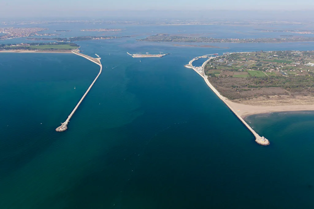An aerial view of two long breakwaters extending from the coastline into the blue sea.