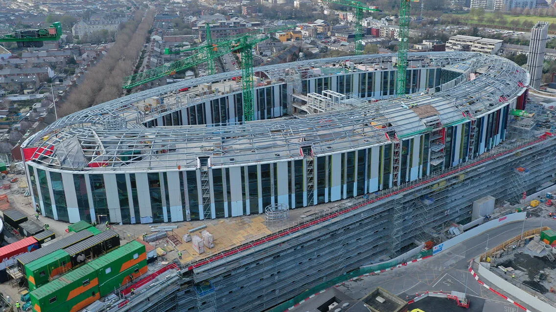 An aerial view of a large, oval-shaped stadium under construction, featuring exposed steel framework for the roof and a facade of vertical panels.