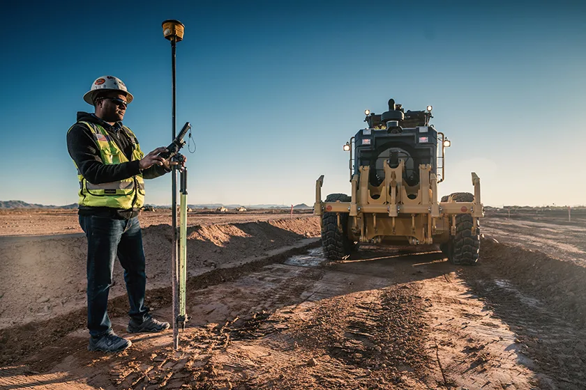 Surveyor uses GPS equipment at a construction site with a grader in the background.