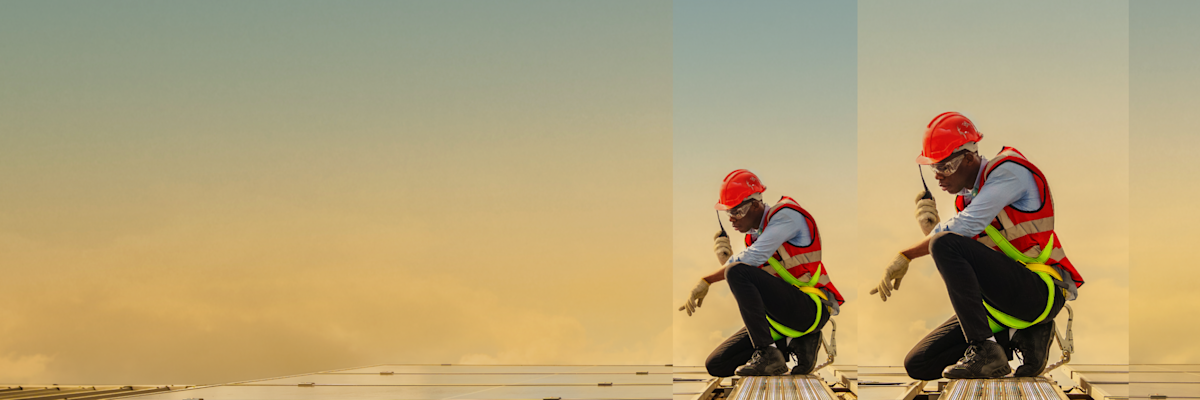 A man holding a radio, wearing a hard hat and orange hi-vis vest, is kneeling on a roof and pointing at solar panels.