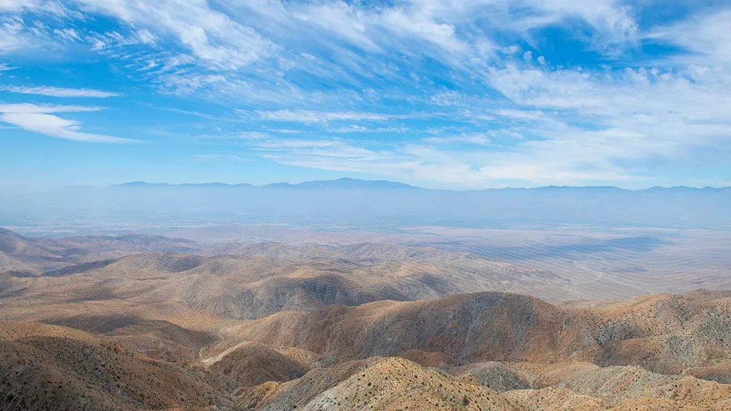 High-angle panoramic view of desert hills and the Coachella Valley from Keys View in Joshua Tree National Park under a blue sky with wispy clouds.