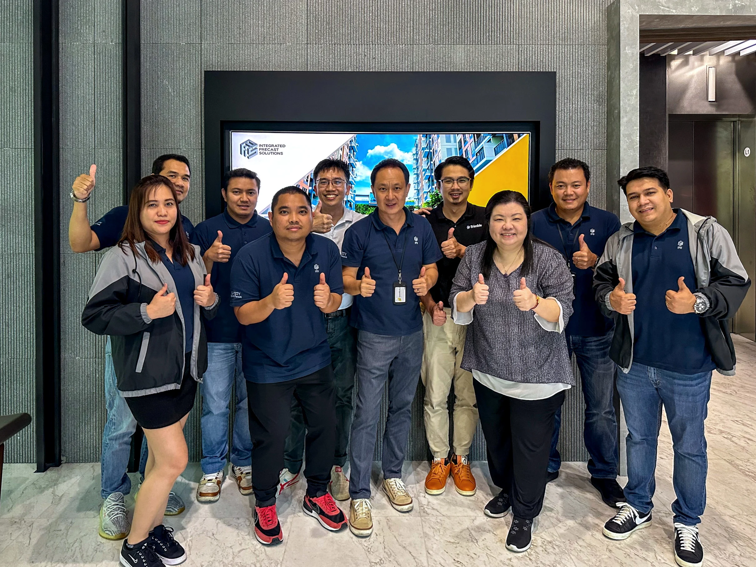 A group of people in matching navy blue shirts and jackets stand in front of a large digital display and an elevator, all giving a thumbs up.