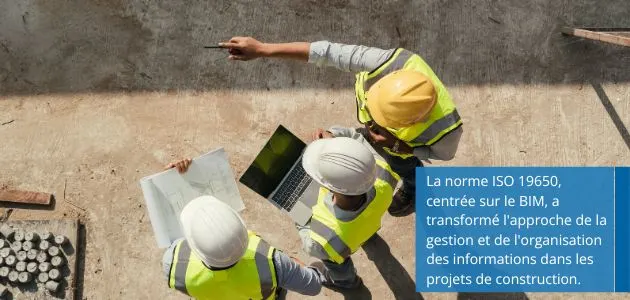 Aerial view of three construction professionals, wearing yellow vests and hard hats, collaborating on blueprints and a laptop at a construction site.