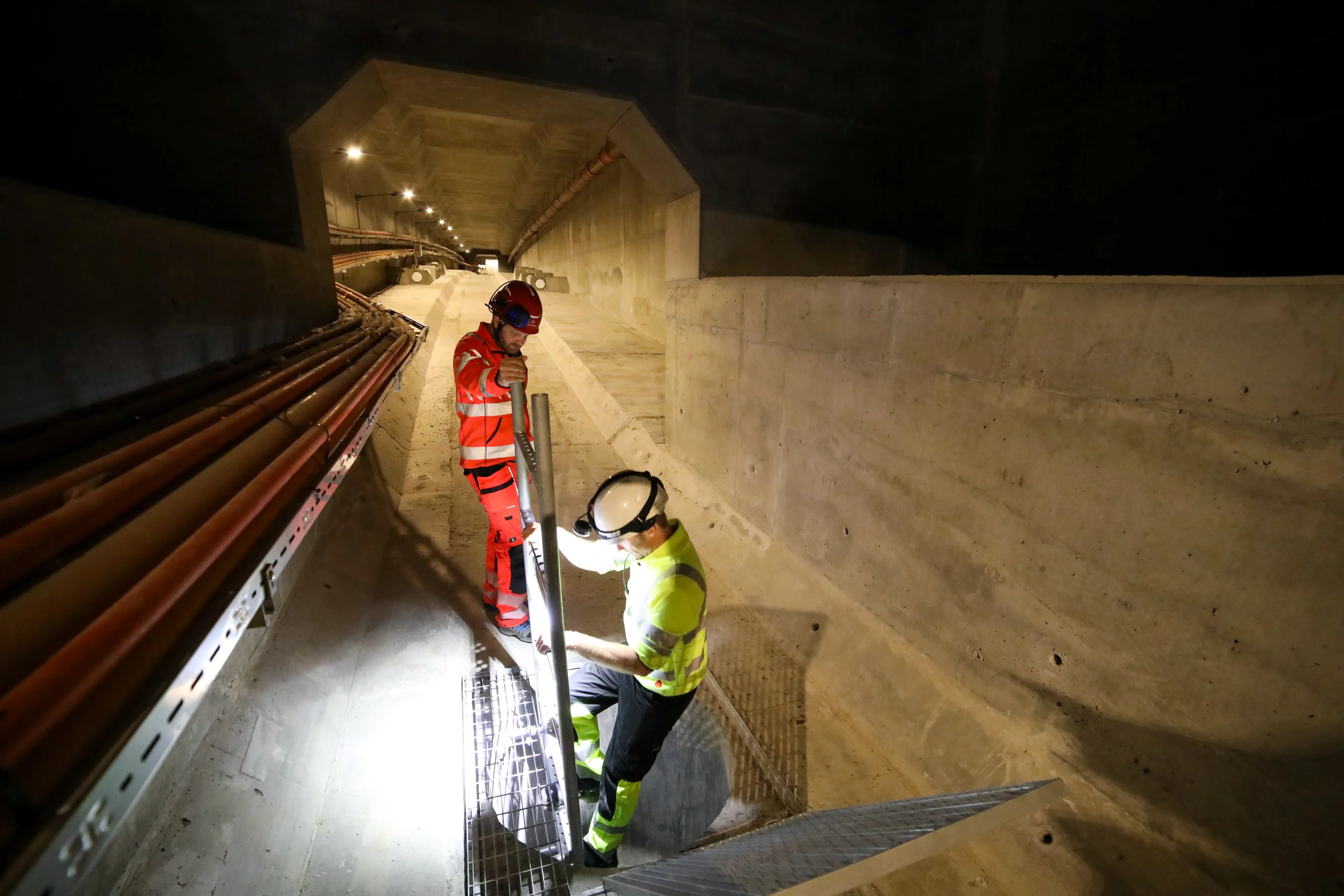 Two workers in hard hats and safety vests inspect infrastructure in a concrete tunnel.
