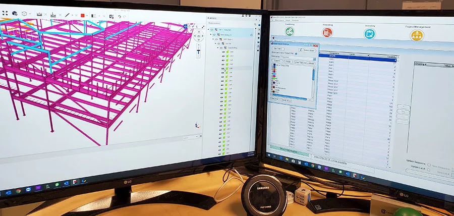 Two monitors display architectural designs, one has a 3D model of a pink and blue steel building structure and the other showing data tables & graphs.