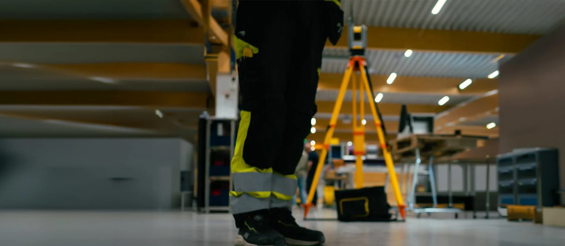 Inside a warehouse, a man in safety gear stands in the foreground, with a Trimble Ri total station in the background.