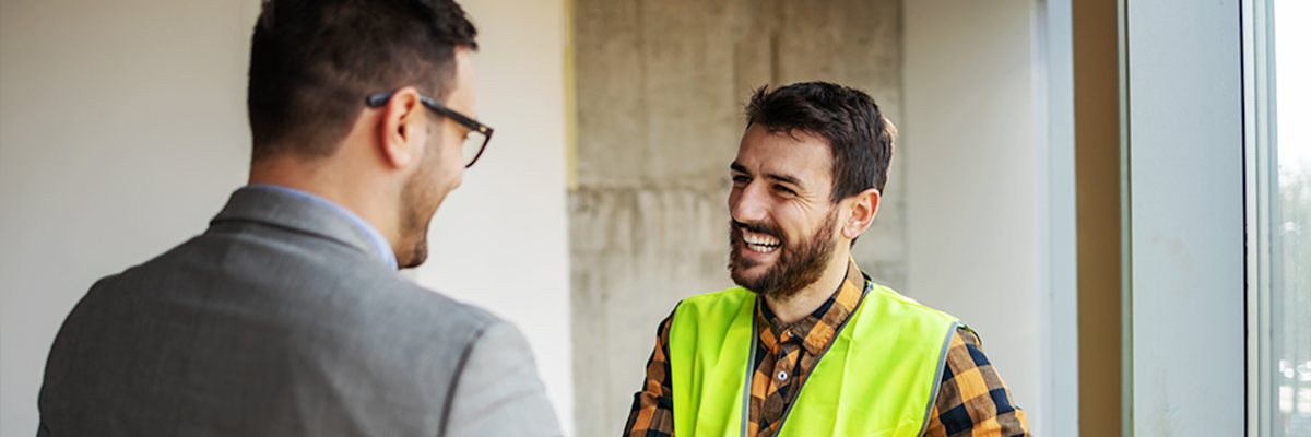 Two men conversing in a construction site, one wearing a bright yellow safety vest and plaid shirt, the other in a gray suit.