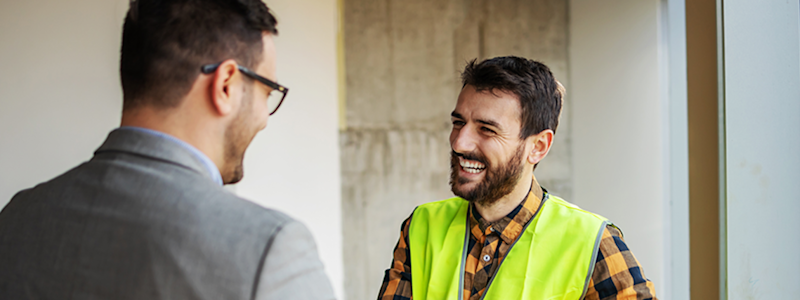 Two men conversing in a construction site, one wearing a bright yellow safety vest and plaid shirt, the other in a gray suit.