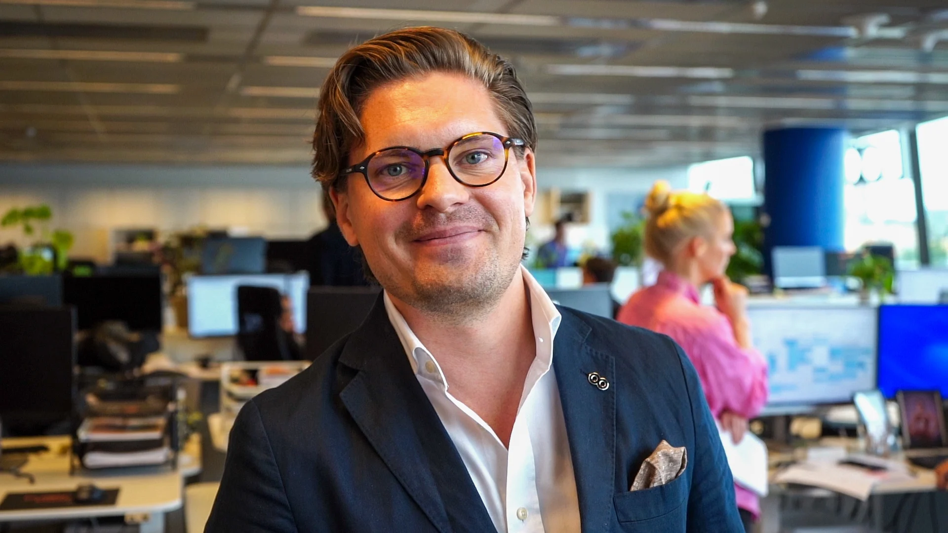 Erik, a man with blond hair and glasses, smiles at the camera in a dark suit jacket.