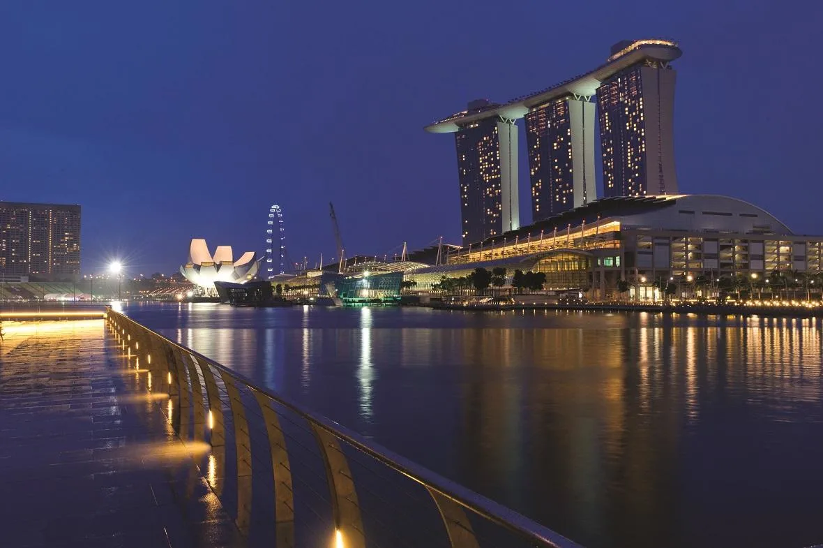 The Marina Bay Sands hotel complex is illuminated at night, reflecting in the calm water, with the Singapore Flyer and other city buildings.