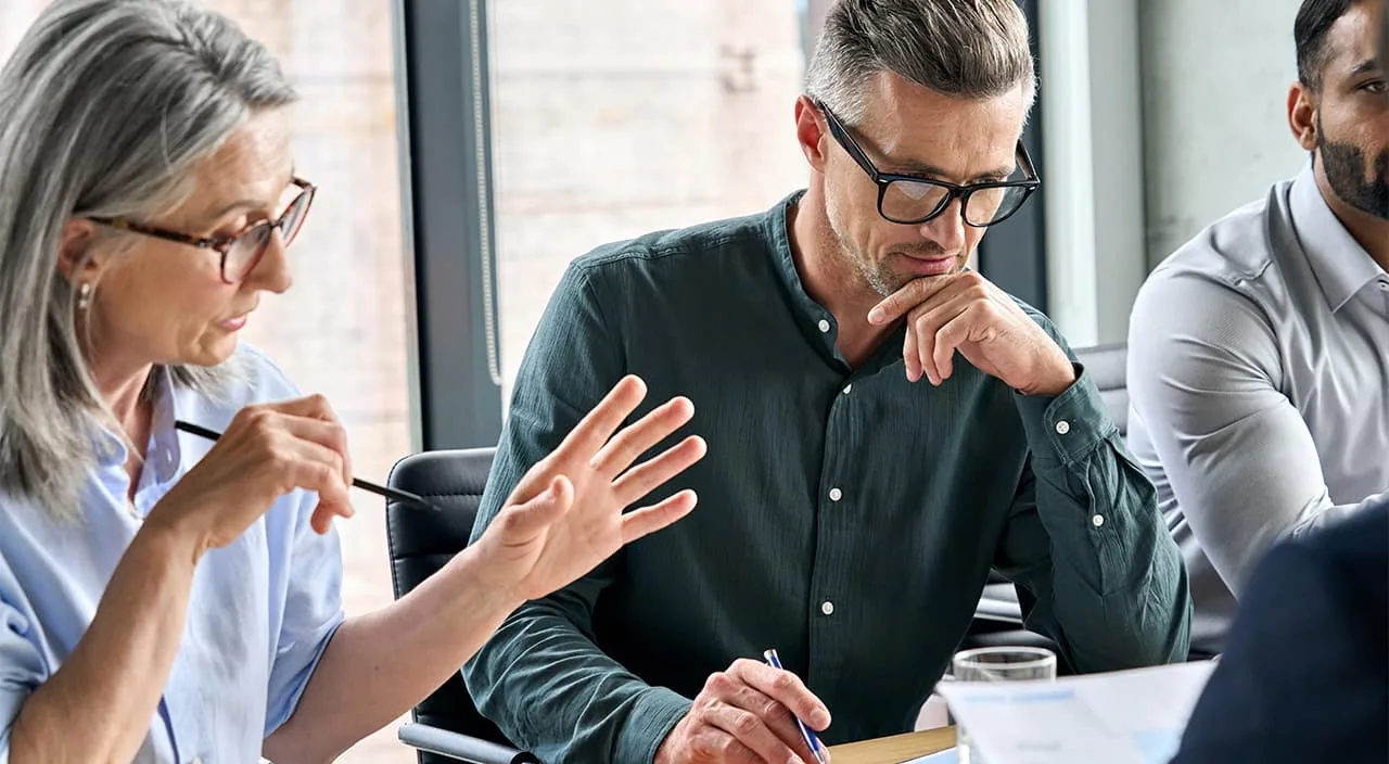 Business meeting: woman speaks and gestures with a pencil, man in glasses listens intently with a pen.