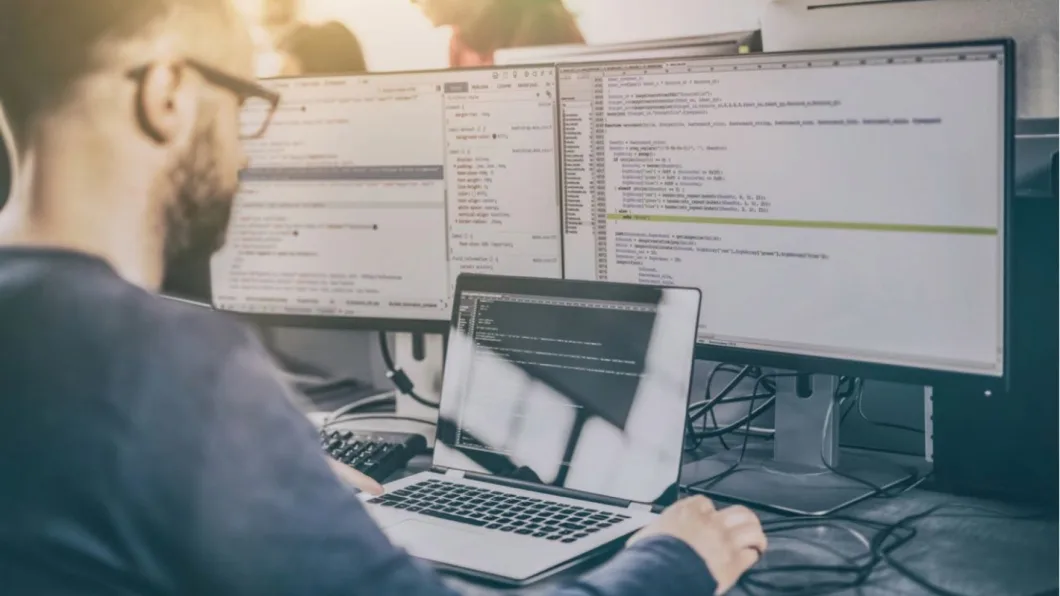 A person works at a desk with a laptop and multiple monitors displaying code, in a modern, well-lit tech workspace.