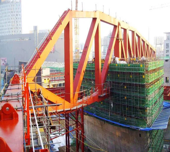 Large orange trusses frame an arched section of a building under construction, supported by green scaffolding.