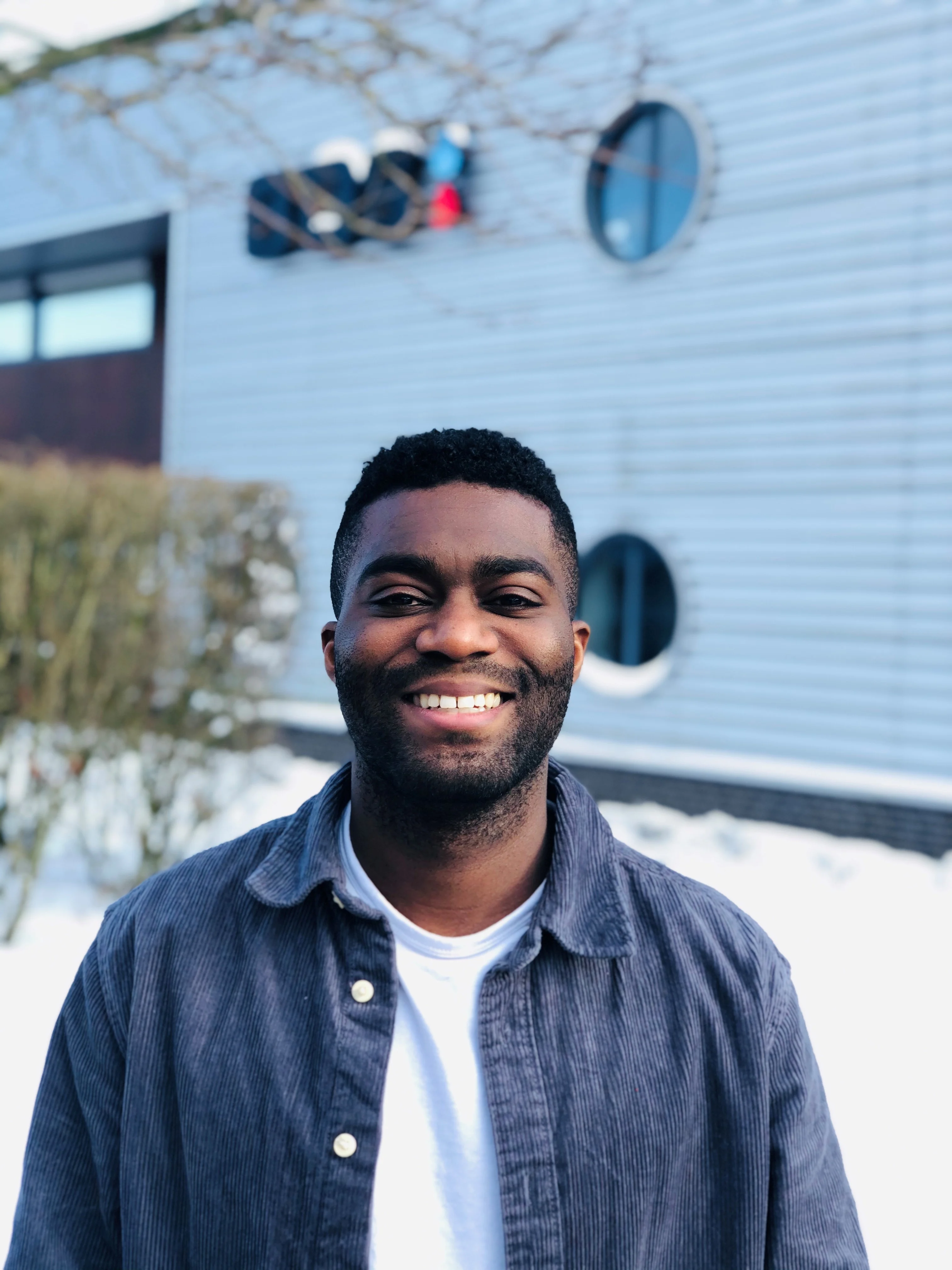 Smiling Black man in a blue corduroy shirt, outdoors in winter with a blurry building background.
