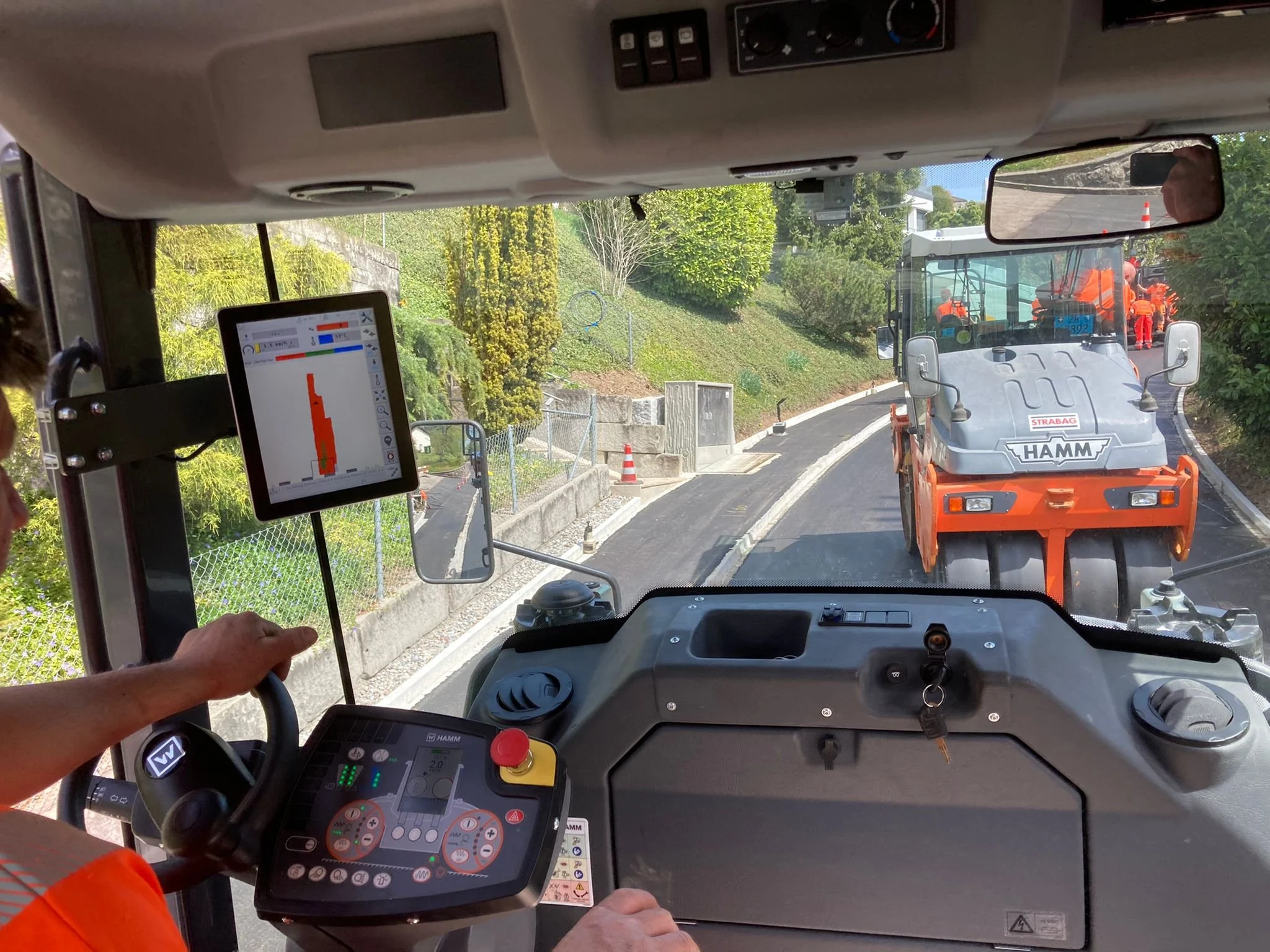View from inside a road roller cabin with control panel and screen, with another roller and workers on a freshly paved road.