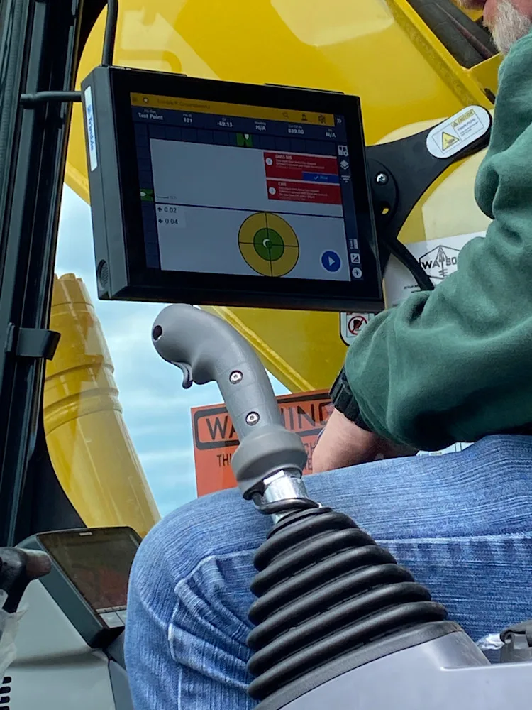 Operator uses a touchscreen display and joystick control in a yellow excavator cab.