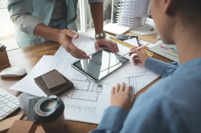 Two designers discuss architectural plans on a table with a tablet, blueprints, and material samples.