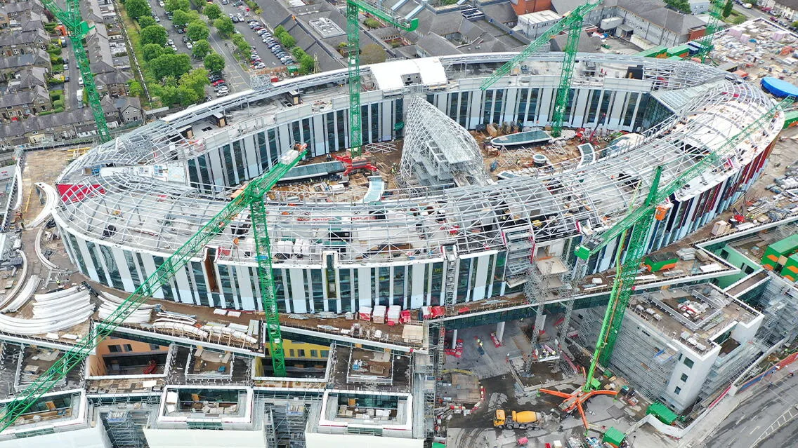 Aerial view of the St. James's Hospital structure under construction in Dublin, with multiple green cranes surrounding the oval-shaped building.