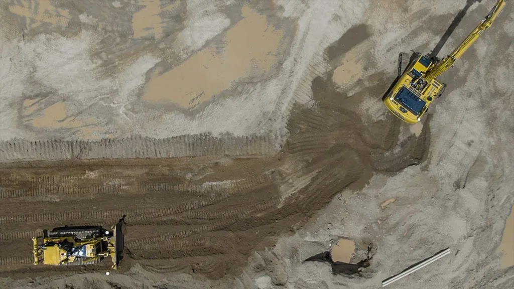 Aerial view of a construction site with a yellow excavator and bulldozer on muddy ground.