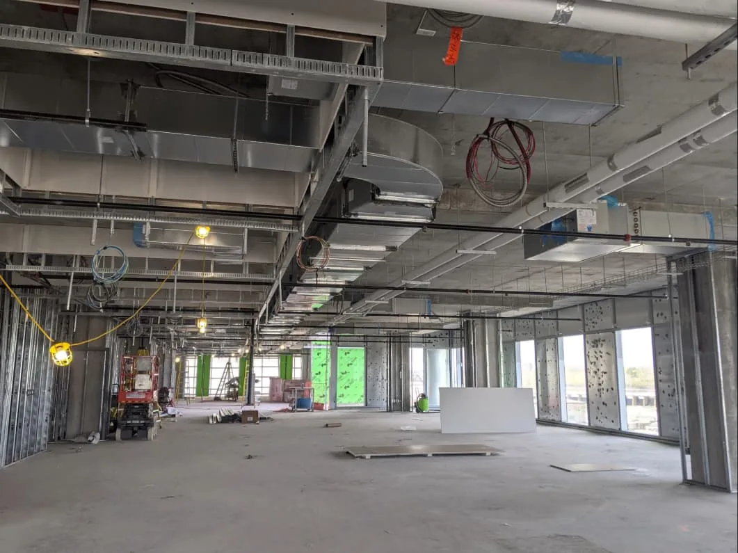 An open room in a construction site shows an open ceiling with visible in-progress electrical and HVAC