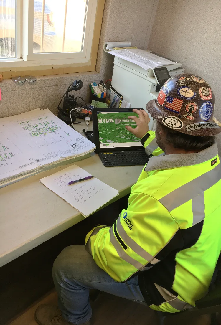A construction worker in a neon yellow jacket and hard hat studies a digital rendering on a laptop, with large stacks of blueprints nearby.