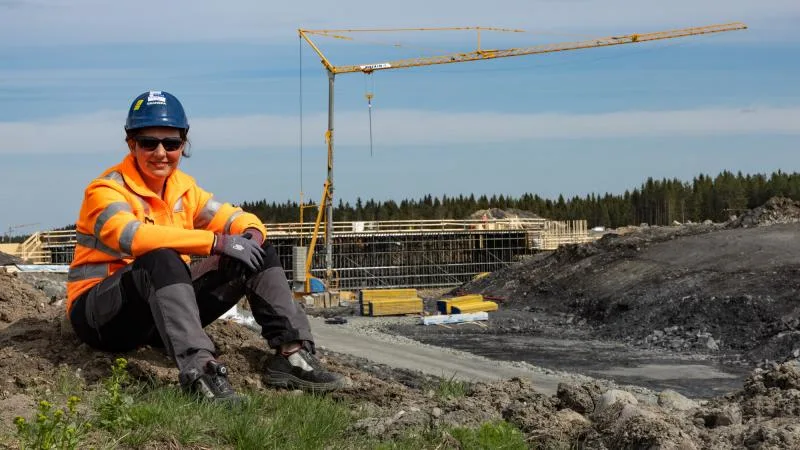 Woman in construction gear sits on a hill at a construction site with a crane.