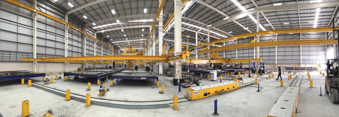 Panoramic view of a modern factory interior with overhead cranes, automated guided vehicles, and large concrete slabs on a manufacturing line.