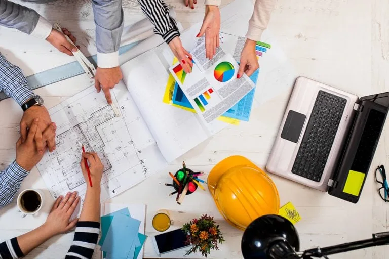 Team collaboration on architectural plans and business charts, viewed from above, with a laptop, hard hat, and desk items.