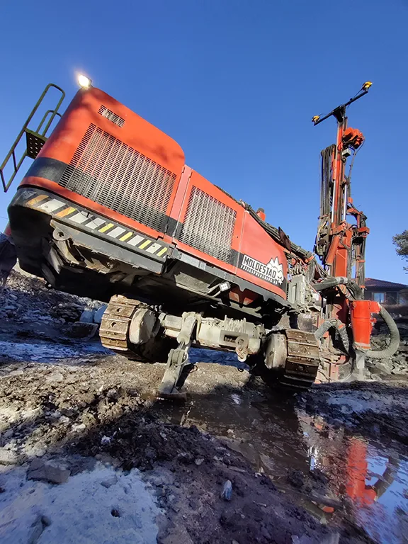 Orange drilling rig with tracks operating in muddy terrain under a clear blue sky.