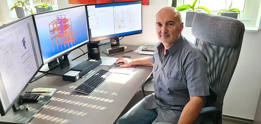 Man at a desk with three computer monitors displaying 3D engineering models and data.