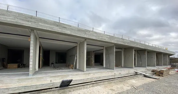 A long, unfinished concrete building with multiple open doorways and a wire fence along the top edge under a cloudy sky.
