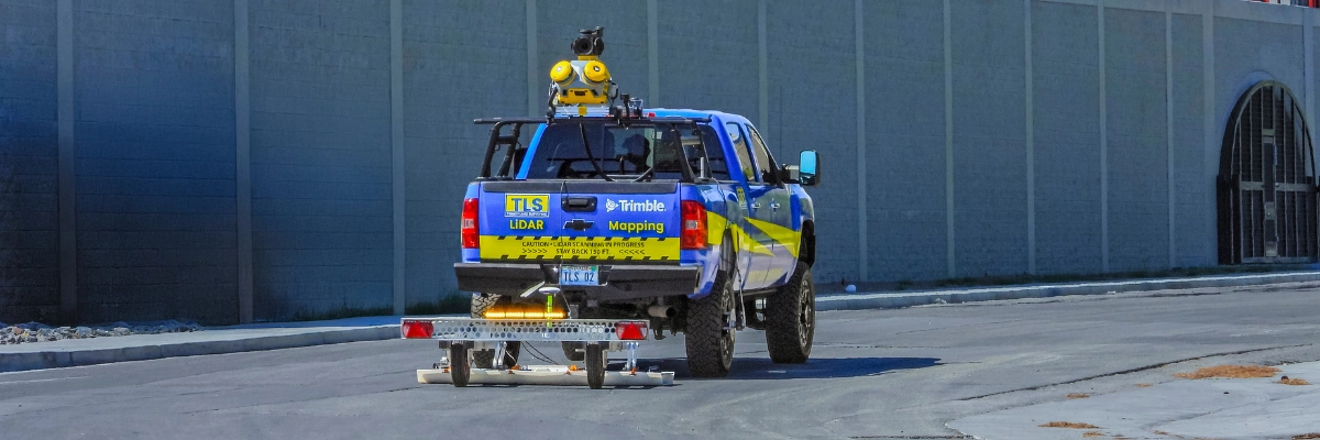 A blue and yellow TLS truck equipped with Trimble LiDAR mapping technology and sensors on the roof, towing a small trailer on a road.
