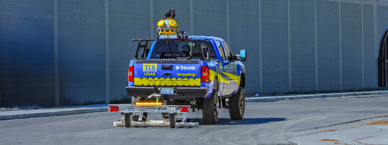 A blue and yellow TLS truck equipped with Trimble LiDAR mapping technology and sensors on the roof, towing a small trailer on a road.