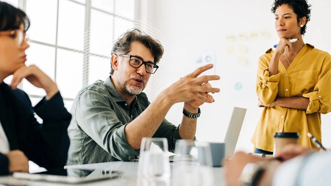 Diverse professional team in a meeting; a man speaks while two women listen thoughtfully in a bright office.
