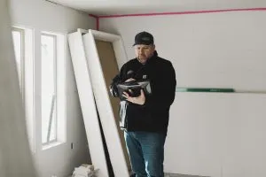 A man in a black jacket and blue jeans holding a black bag in a room under construction.