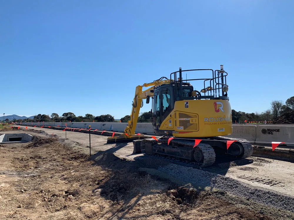 Yellow excavator on a construction site with concrete barriers and orange markers.