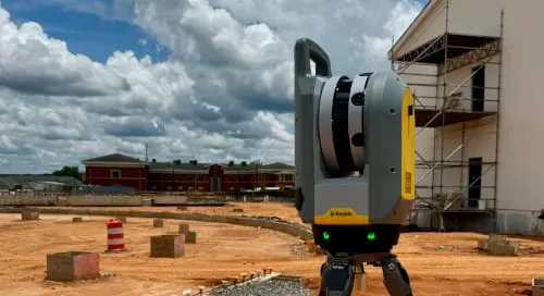 A Trimble surveying instrument on a tripod at a construction site with scaffolding, dirt ground, and cloudy sky.