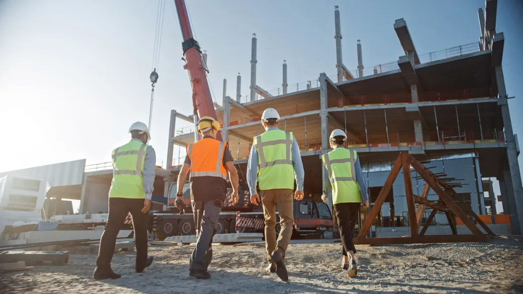 Four people wearing hardhats and safety vests walk toward a building construction site.