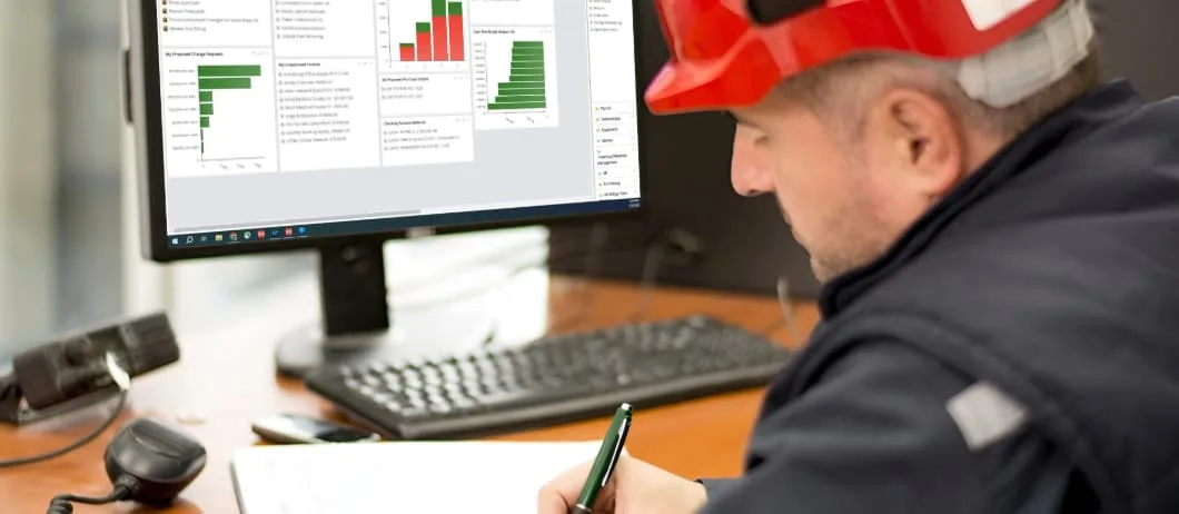 A person wearing a red hard hat sits at a desk, writing with a pen while looking at a computer monitor.