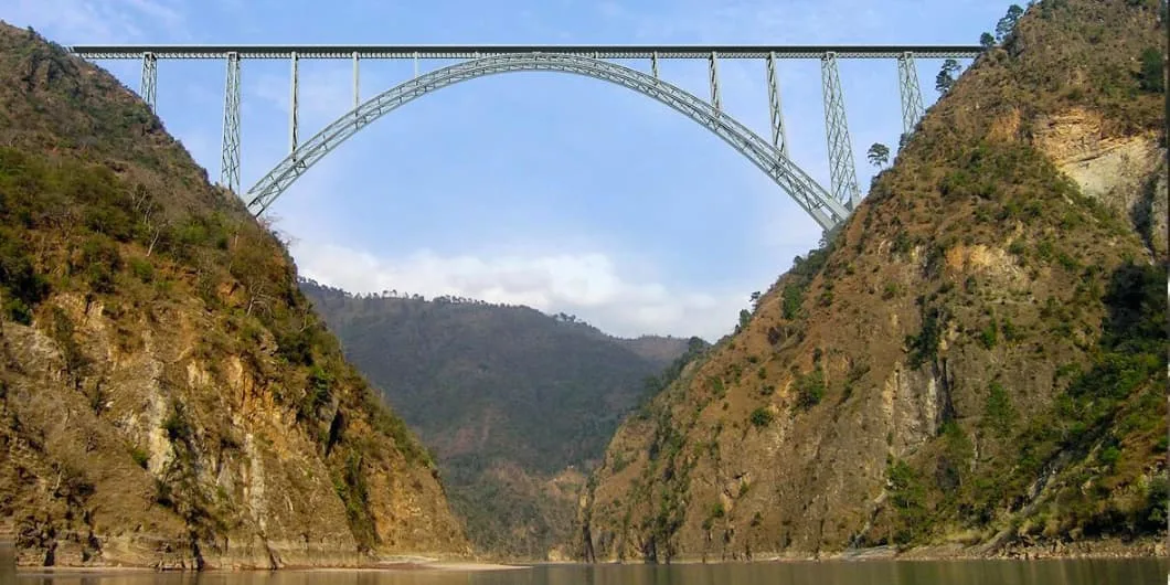 A steel truss arch bridge spans a wide river valley between two steep, rocky mountainsides.