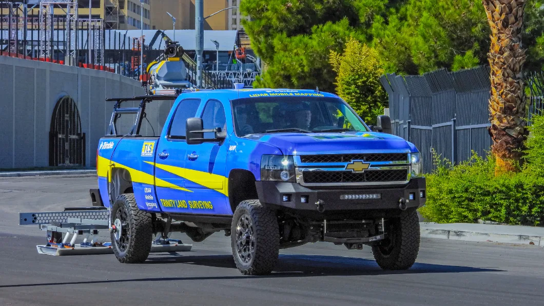 Blue and yellow Chevrolet truck branded Trinity Land Surveying with LiDAR mobile mapping equipment on roof, driving on a road.