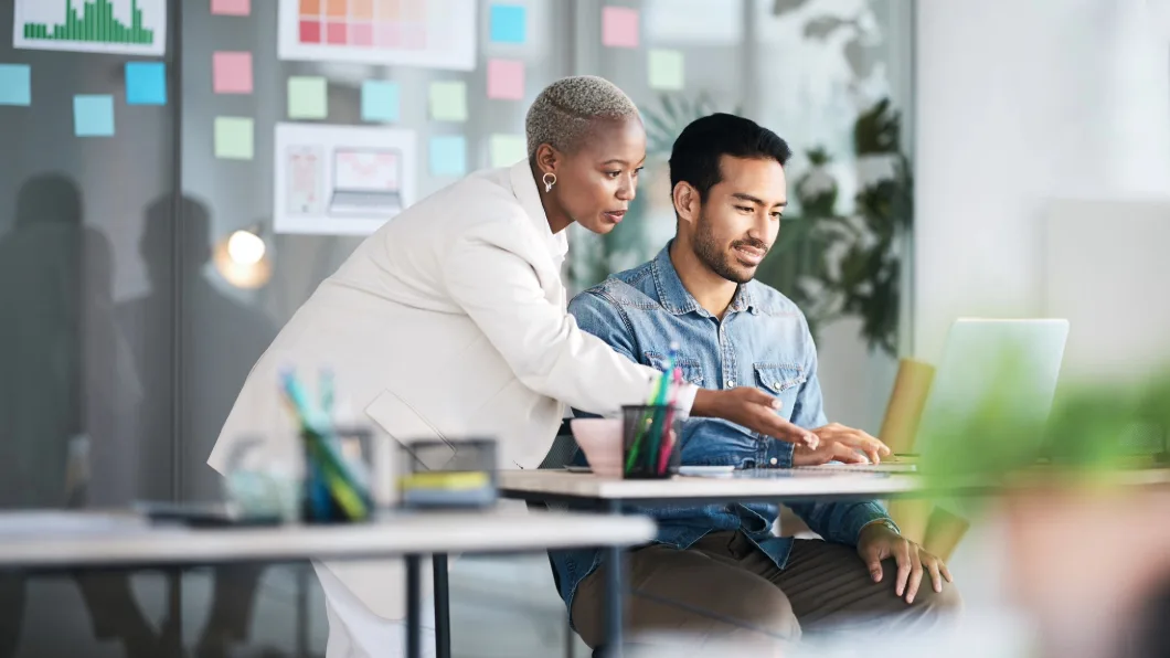 Colleagues collaborating at a desk with a laptop and sticky notes on the wall.