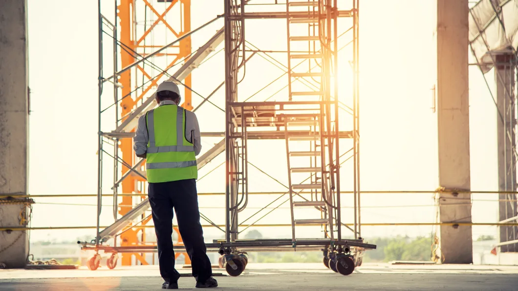Construction worker in safety gear stands near scaffolding at a building site during sunset.