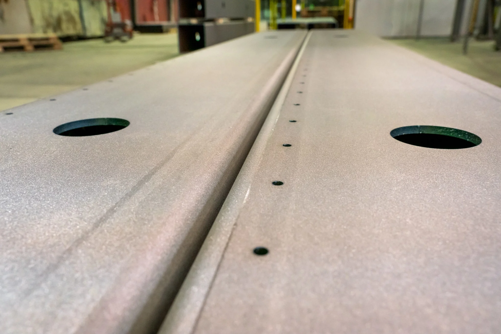 Large metal components with circular holes and evenly spaced small holes lie on the floor of a manufacturing facility.