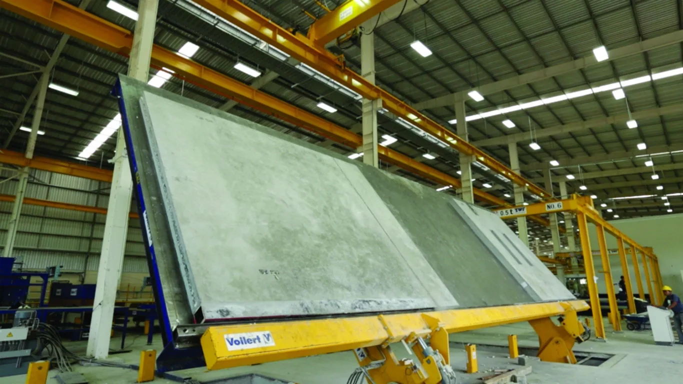 A large, gray precast concrete panel is shown on a yellow tilting table in a factory setting.