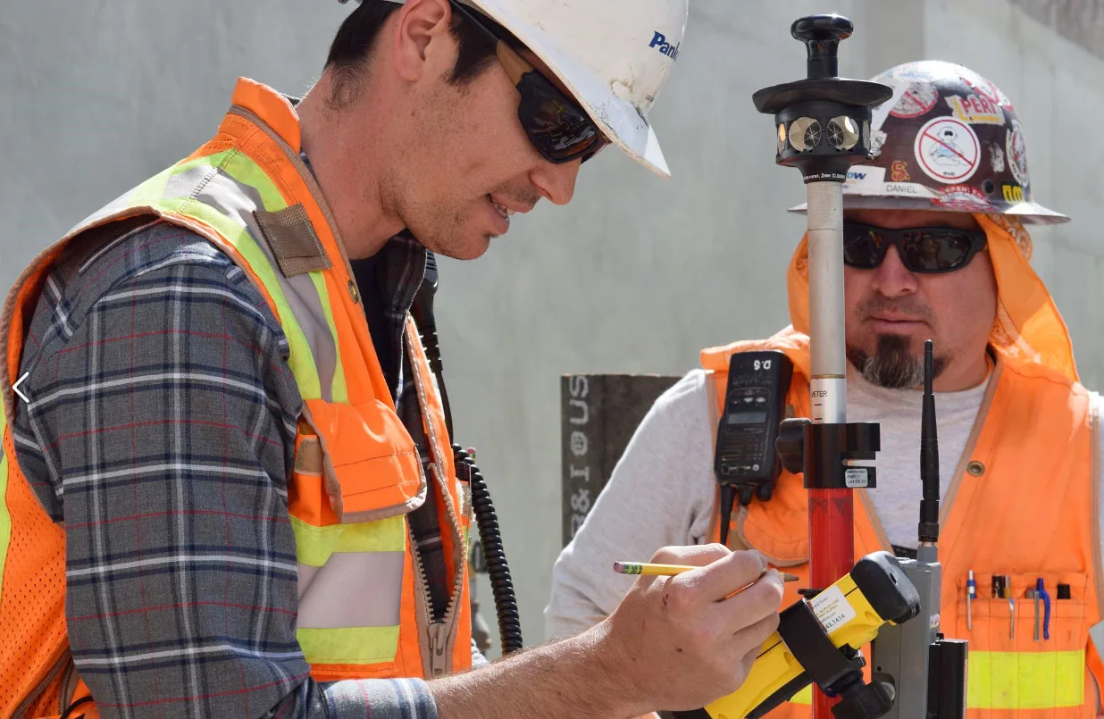 Two construction workers in hard hats and safety vests examine surveying equipment on a job site.