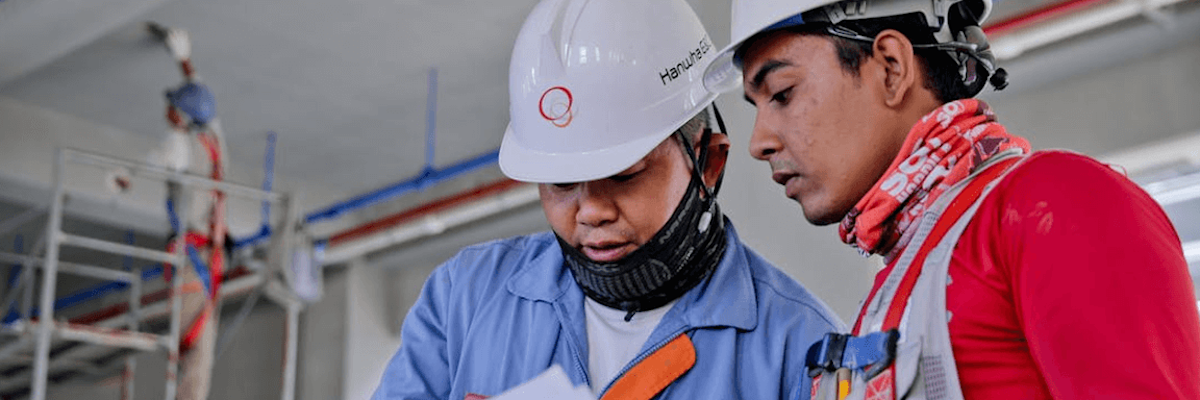 Two construction workers in safety gear review documents indoors, with a third worker on scaffolding in the background.