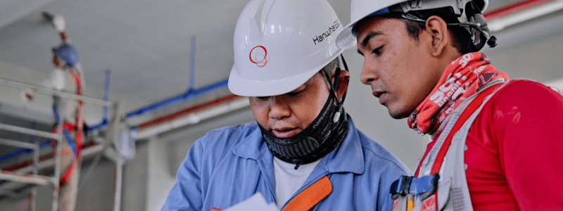 Two construction workers in safety gear review documents indoors, with a third worker on scaffolding in the background.