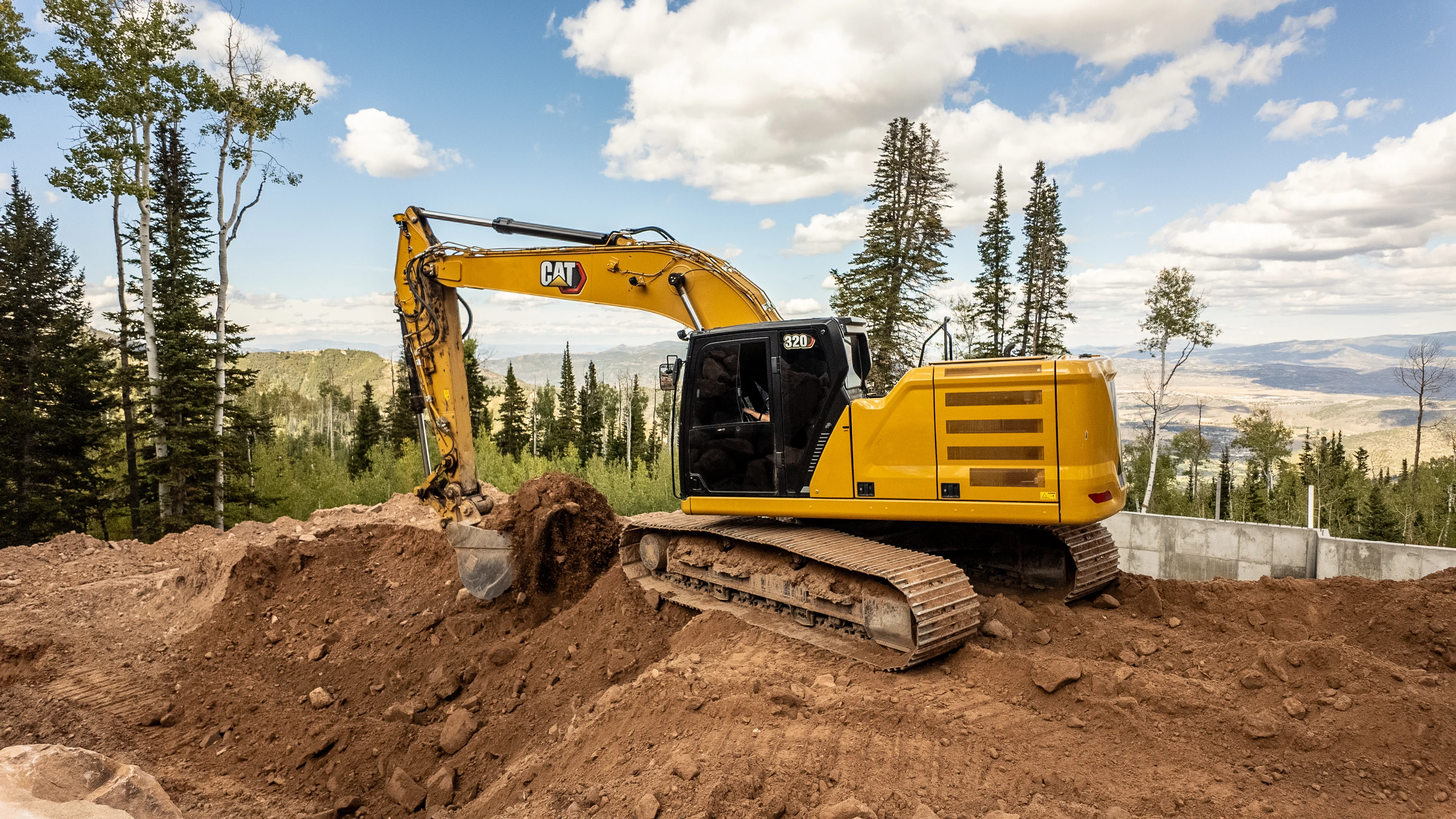 A yellow CAT 320 excavator digs into a pile of red dirt on a mountain construction site, surrounded by trees and a valley view.