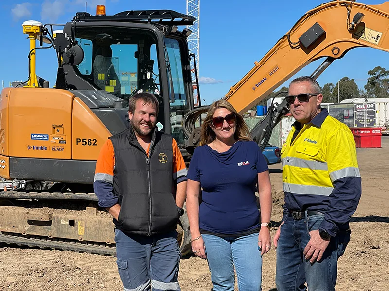Three construction workers stand in front of a large excavator on a sunny day.